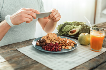 Good combination. Attentive smart creative man sitting by the table holding the cellphone and making photo of food.