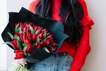 Young woman in a red sweater holding beautiful fresh blossoming red tulips flower bouquet wrapped in a black paper