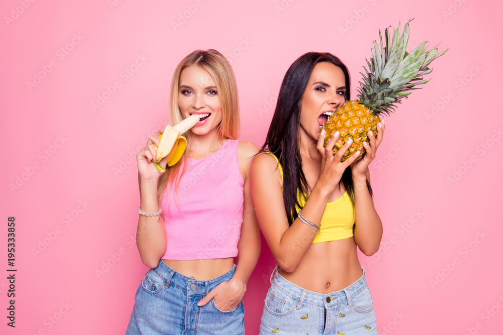 Sticker Carelessness, recreation, rest, relaxation concept. Pretty beautiful cheerful fancy girlfriends wearing clothes and bright tops bite and eating tropical fruit isolated on bright pink background