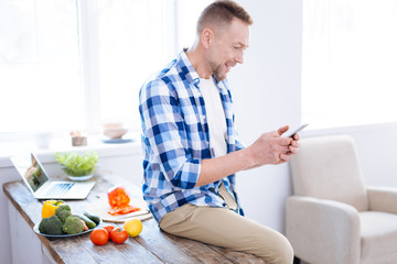 Biohackers recipes. Joyful merry jolly man using phone for looking through article about biohacking and sitting on surface