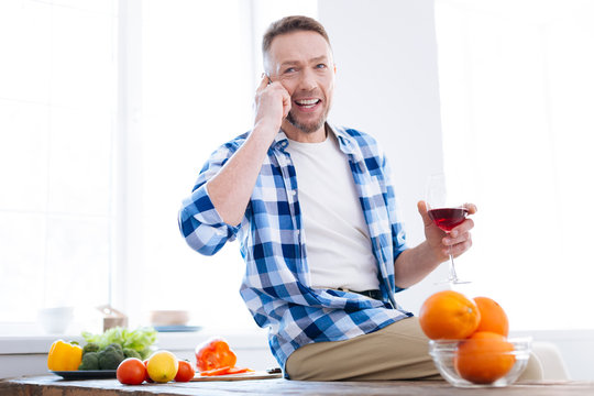 Light meal. Pleased talkative nice man using phone while holding glass of wine and sitting on surface