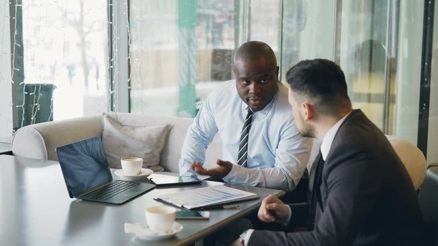 Two multi-ethnic businessmen looking at diagrams and graphs on laptop screen and discussing the financial report of their startup in modern cafe during lunch time.