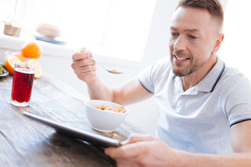 Easy to make. Jolly pleasant calm man smiling while staring at tablet screen and tasting granola