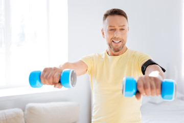 Key trend. Jovial jolly handsome man using dumbbells while exercising and looking at hand