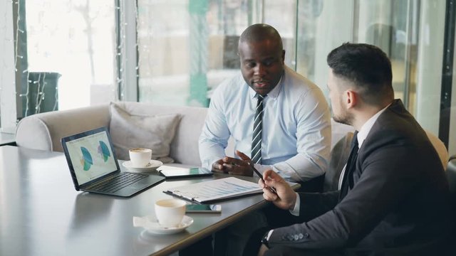 Two confident businessmen looking at diagrams and graphs on laptop computer and discussing the financial report and statistics of their business project in modern cafe during lunch time.