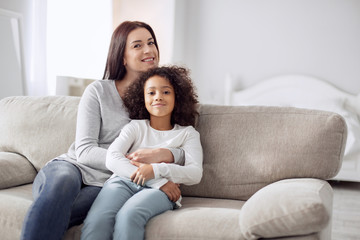 I feel happy. Beautiful cheerful young dark-haired woman smiling and hugging her daughter while sitting on the couch