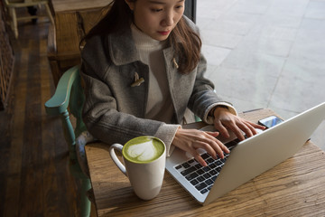 Fototapeta premium Beautiful young woman's hands busy working on her laptop sitting at wooden table in a coffee shop. And closeup of Matcha Latte.