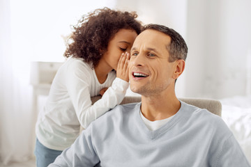 My secrets. Good-looking happy dark-haired father sitting in the arm-chair and smiling and his daughter whispering in his ear