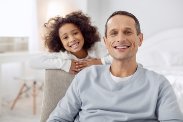 Obraz premium My precious. Handsome exuberant dark-haired father sitting in the arm-chair and smiling and his daughter standing behind him