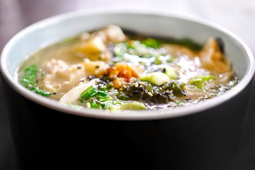 Mushroom chicken Wonton soup on a black plate and on a wooden table. Chinese food. On the table are flowers in a transparent vase and a lilac pot with green leaves. Top view with copy space. 