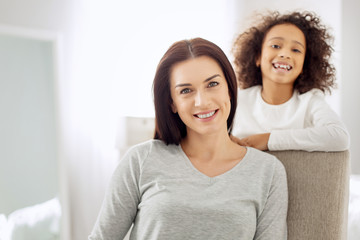 Glad woman. Beautiful alert dark-haired woman sitting in the arm-chair and smiling and her daughter standing behind her