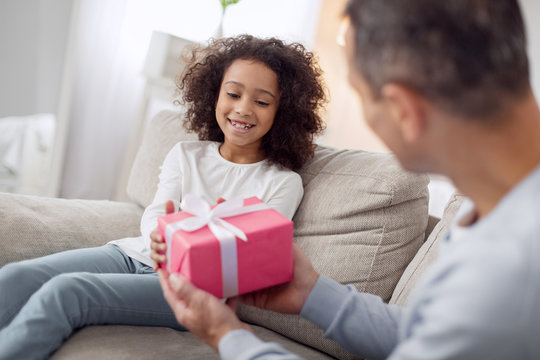 Happy Birthday. Nice joyful curly-haired girl sitting on the sofa and smiling and her daddy giving her a gift