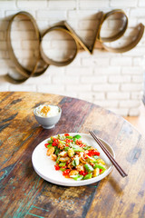 Chicken kung pao on a white plate and on a light wooden table. Chinese food. Food sticks. On the back of a blurry background, a white brick pad is covered with an word  love. Side view with copy space