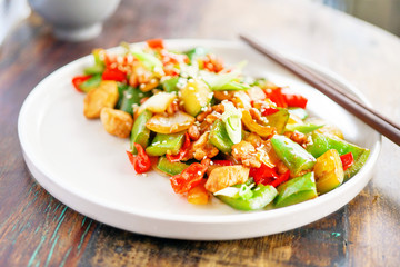 Chicken kong pao on a white plate and on a light wooden table. Chinese food. Food sticks. In the frame, the hand holds the dumpling with chopsticks. Close-up view with copy space. 