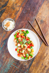 Chicken kong pao on a white plate and on a light wooden table. Chinese food. Food sticks. In the frame, the hand holds the dumpling with chopsticks. Top view with copy space. 