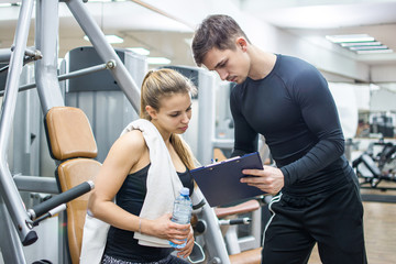 Young sporty woman and her personal trainer looking together at sports results on clipboard in gym. - obrazy, fototapety, plakaty