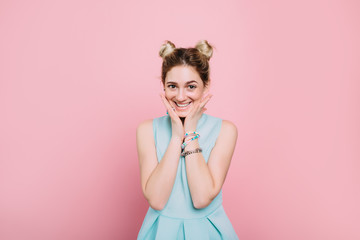 happy facial expression. young trendy woman wearing turquoise dress making a surprised face and looking at camera while standing against pastel pink background