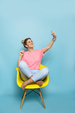 Fashionable young woman making selfie, sitting on a yellow chair, against a blue pastel background . youth lifestyle