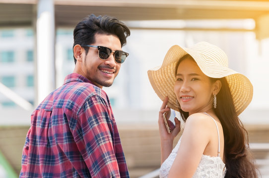 Young Asian couple lover be happy during honeymoon trip in big city, Asian man smiling wearing sun glasses with casual cloth, woman wearing white dress and big hat.