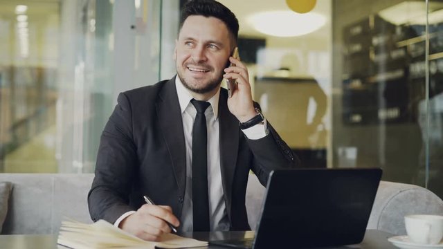 Happy bearded businessman in black business suit sitting at the table, smiling and talking on mobile phone in modern cafe