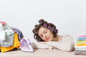 Obraz na płótnie Canvas Tired housewife with curlers on hair in light clothes fell asleep on family clothing on ironing board with iron. Woman isolated on white background. Housekeeping concept. Copy space for advertisement.