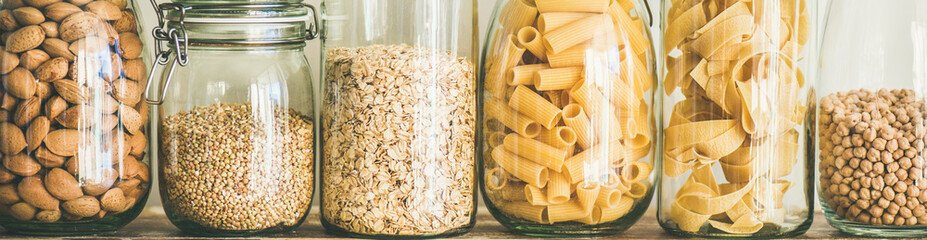 Various uncooked cereals, grains, beans and pasta for healthy cooking in glass jars on wooden table, white background, close-up, wide composition. Clean eating, vegan, balanced dieting food concept