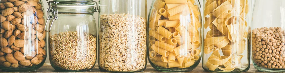 Verschiedene ungekochte Cerealien, Körner, Bohnen und Nudeln für gesundes Kochen in Glasgefäßen auf Holztisch, weißer Hintergrund, Nahaufnahme, breite Komposition. Sauberes Essen, veganes, ausgewogenes Ernährungskonzept © sonyakamoz