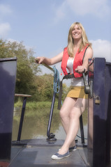 Woman wearing a life jacket and using the tiller to steer a narrowboat along a canal in the UK