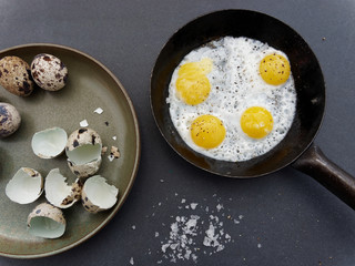 Wachtelspiegeleier in altem Pfännchen, Wachteleier und Eierschalen auf grünem Steingutteller, grobes Salz, auf Schieferplatte, im Anschnitt