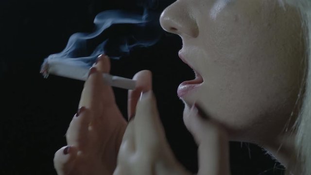 Close up of a depressed blonde teenager girl sitting in the dark lightning a weed cigarette inhaling and exhaling clouds of smoke