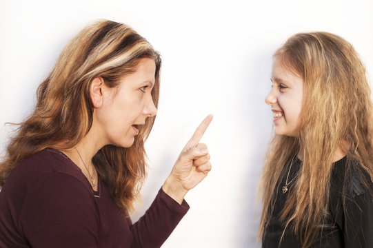 Mom Scolding Her Daughter With An Angry Face And Waves Her Finger On A White Background. Daughter At The Same Time Just Smiles