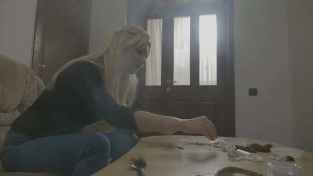 Beautiful young blonde woman rolling a marijuana cigarette at home and smoking it to calm down after being fired from work