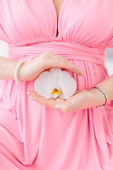 Young beautiful pregnant girl in a pink dress is ssitting on the bed with orchid in her hand