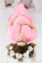 Fototapeta na wymiar Young beautiful pregnant girl in a pink dress lying in the bed with orchid in her hair