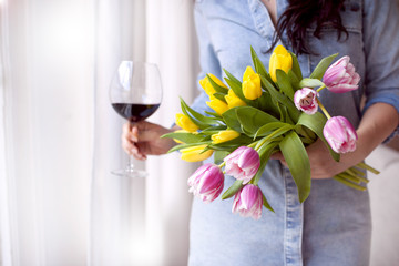 A woman with a glass of red wine, holding a bouquet of fresh tulips of yellow and pink in her hands. A gift for the beloved. Near the window. Postcard