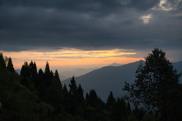 Series of Himalayan mountains in the twilight similar to the sea waves go away against the backdrop of silhouettes of the forest in the foreground.