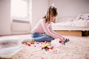 Obraz na płótnie Canvas Beautiful thoughtful little toddler girl sitting on the carpet of her bright room and playing with plastic toys.