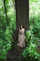young woman in vintage dress pressed against the trunk of a huge forest tree