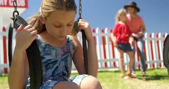 Upset girl sitting on a swing while friends talking in the background 