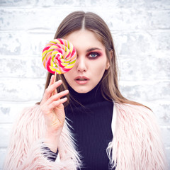 A girl with lollipop. Girl in casual clothes. Brick wall background. Gloomy makeup. Fashionable woman. Casual wear. Trend clothing. Toned image.