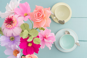 Crepe paper flower bouquet with cosmos, roses, echinacea and eucalyptus in a vase on light blue table with coffee cups shot from above