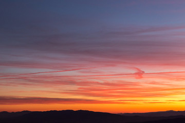 Fototapeta premium Clouds and jet vapor trails creating beautiful, colorful texture in the sky at sunset with mountain profiles on the low part of the frame