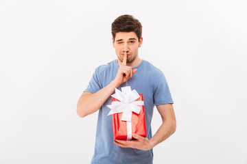 Photo of handsome man holding birthday gift and asking to keep secret with finger on lips, isolated over white wall