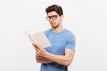 Portrait of young smart man in blue shirt wearing eyeglasses reading book with concentration, isolated over white background