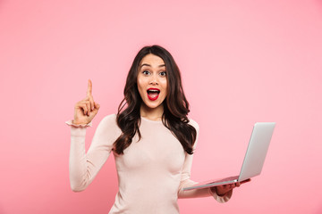 Portrait of agitated female with long dark hair holding silver laptop and pointing finger upward on...