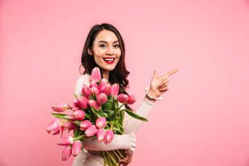 Charming brunette female holding amazing bunch of blossom tulips on women's day and pointing finger on copyspace, isolated over pink background