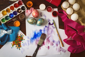 Preprimary painting of Easter Eggs, Brushes, Paints, Eggs, Sweets, Ribbons on the Dark Wooden Table. The concept of Easter. Flat lay, top view