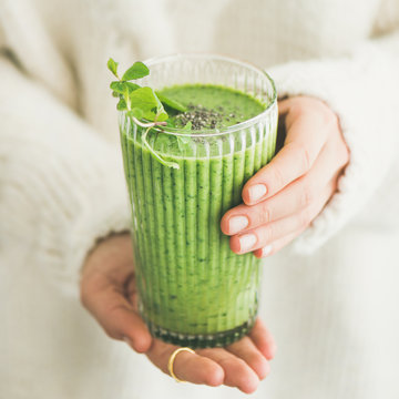 Matcha green vegan smoothie with chia seeds and mint in glass in hands of female wearing white sweater, square crop. Clean eating, detox, alkaline diet, weight loss food concept