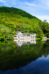 Beautiful abundant natural shades of spring green mountain background mirror reflection on fresh lake Kinrinko with buildings and blue sky, Yufuin
