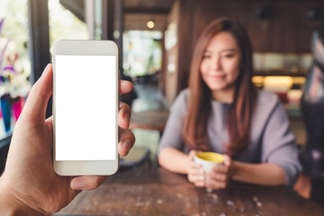 Mockup image of a hand holding white mobile phone with blank desktop screen in cafe with a woman in background